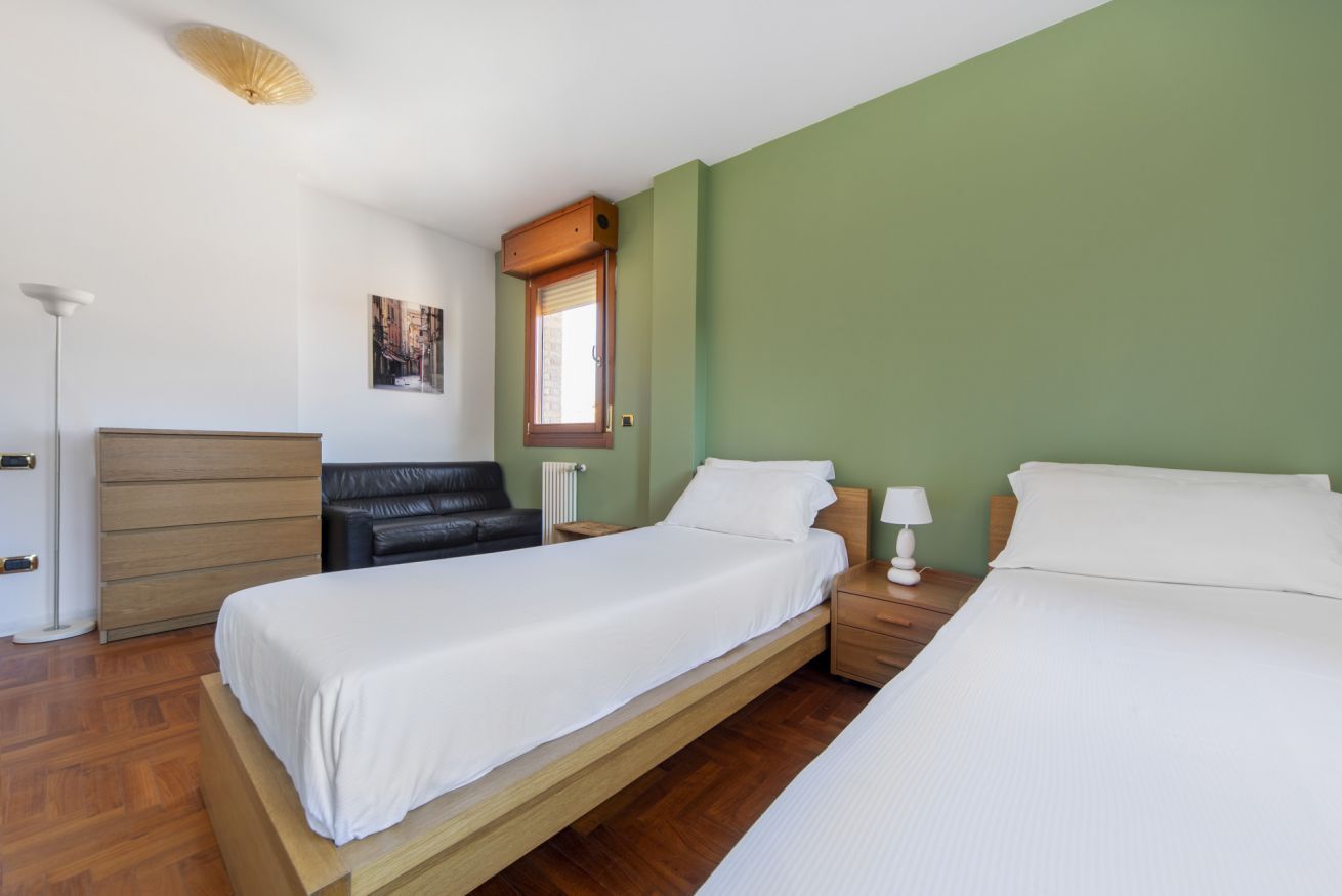 A bedroom with two single beds, a wooden dresser, a black leather sofa, a floor lamp, and a window with blinds.