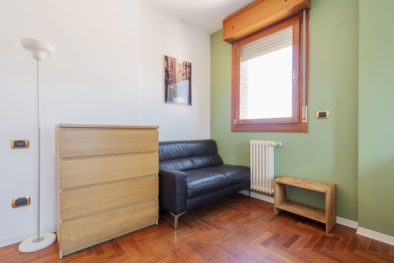 A room with a wooden cabinet, a black leather sofa, a small wooden table, a radiator, and a window with blinds.