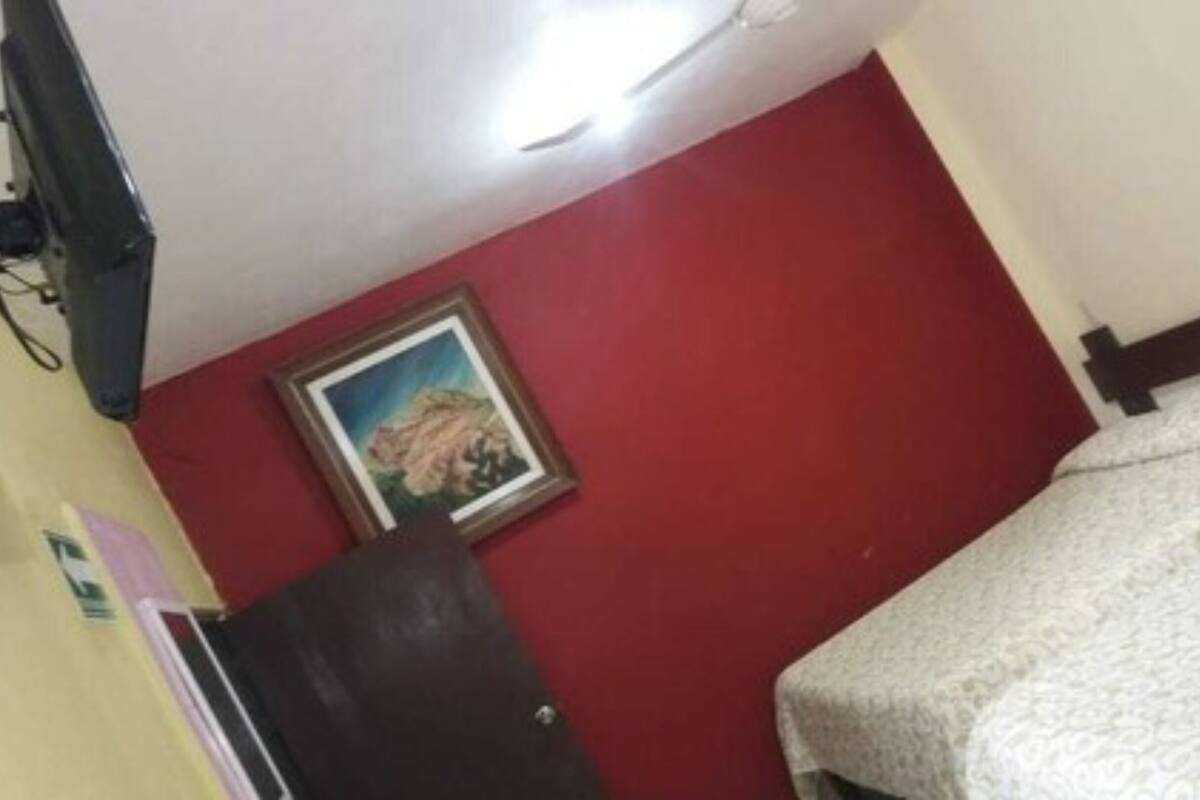 A room with a bed, a red wall, a framed picture, and a television mounted on the wall.