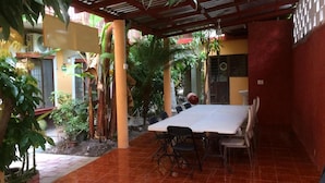 Eine Terrasse im Freien mit einem langen Tisch und Stühlen, umgeben von Topfpflanzen und einem gefliesten Boden.