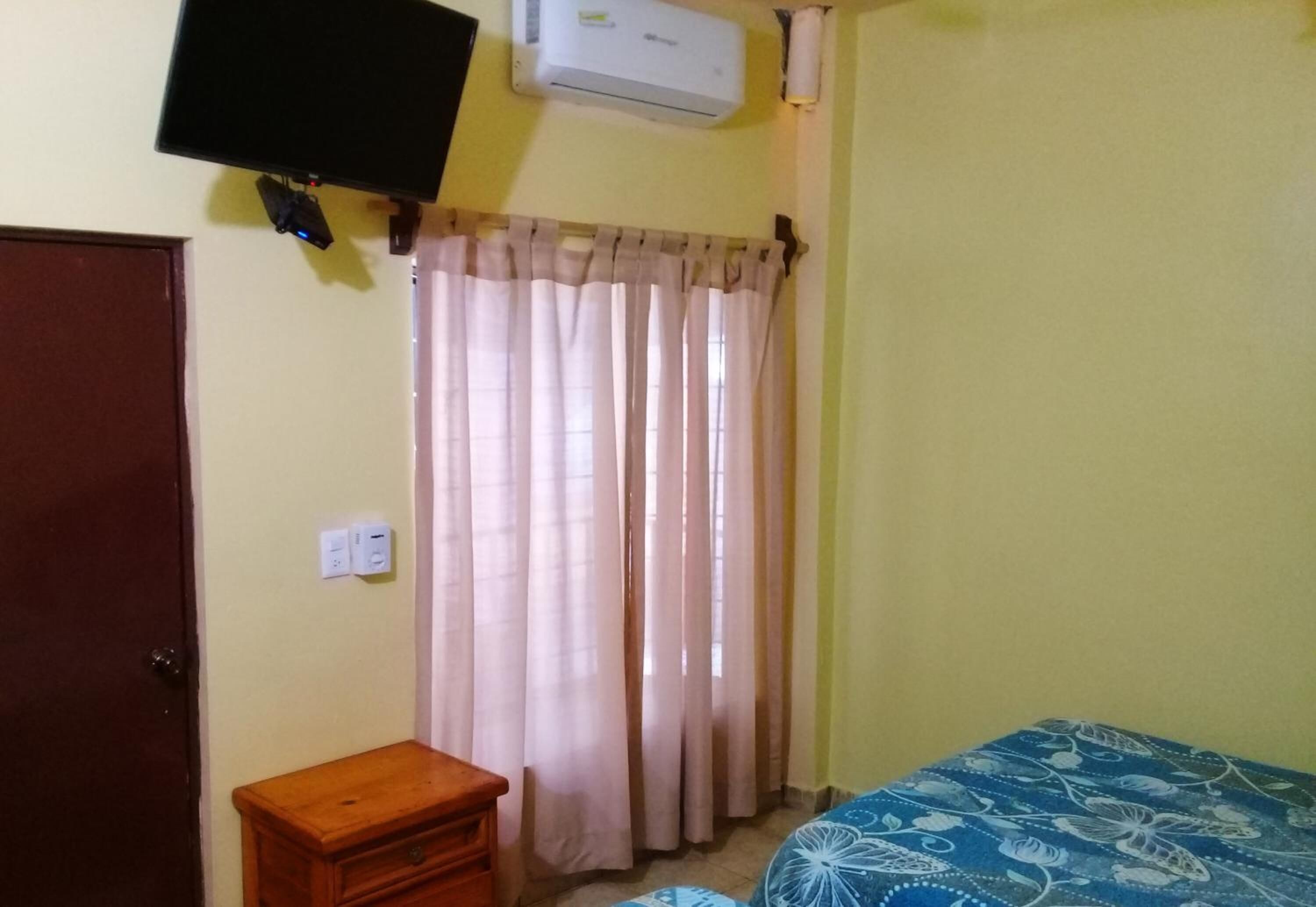 A room with a bed, a small wooden nightstand, a TV mounted on the wall, and a window with curtains.
