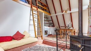 A cozy loft bedroom with a loft bed, a wooden ladder, a fireplace, and a kitchen area in the background. - Junction Retreat - Ohakune Holiday Home (Ohakune)