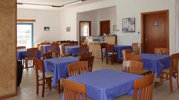 Un'area ristorazione con tavoli rivestiti da tovaglie blu e sedie di legno, una reception e un muro con manifesti informativi.