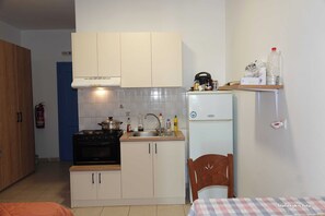Une cuisine de taille réduite, équipée d’une cuisinière, d’un évier et d’un réfrigérateur. On aperçoit une porte bleue au fond, et un extincteur est fixé au mur.