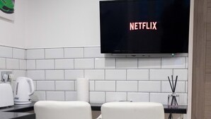 A modern kitchen with two white bar stools, a flat-screen TV displaying 'NETFLIX', and a white tiled backsplash. - Westwood Apartment (Leeds)