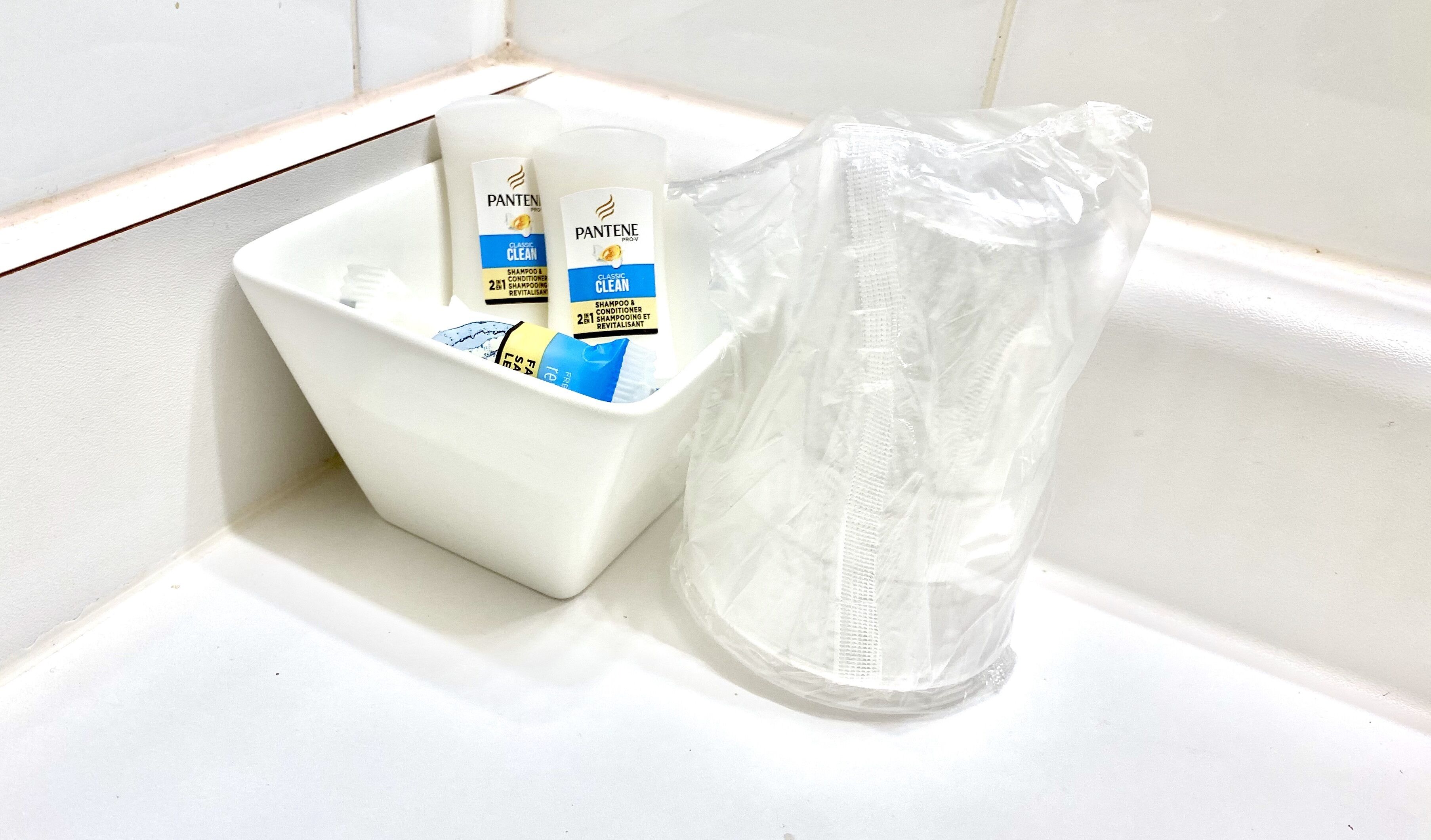 A bathroom corner with a white sink, two bottles of Pantene Classic Clean shampoo and conditioner, and a clear plastic bag.