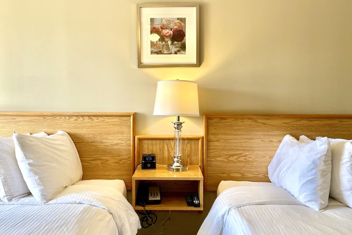 Ein Hotelzimmer mit zwei Betten, einem Nachttisch mit Lampe, einem gerahmten Bild an der Wand und einem kleinen Regal mit Telefon und Fernbedienungen.