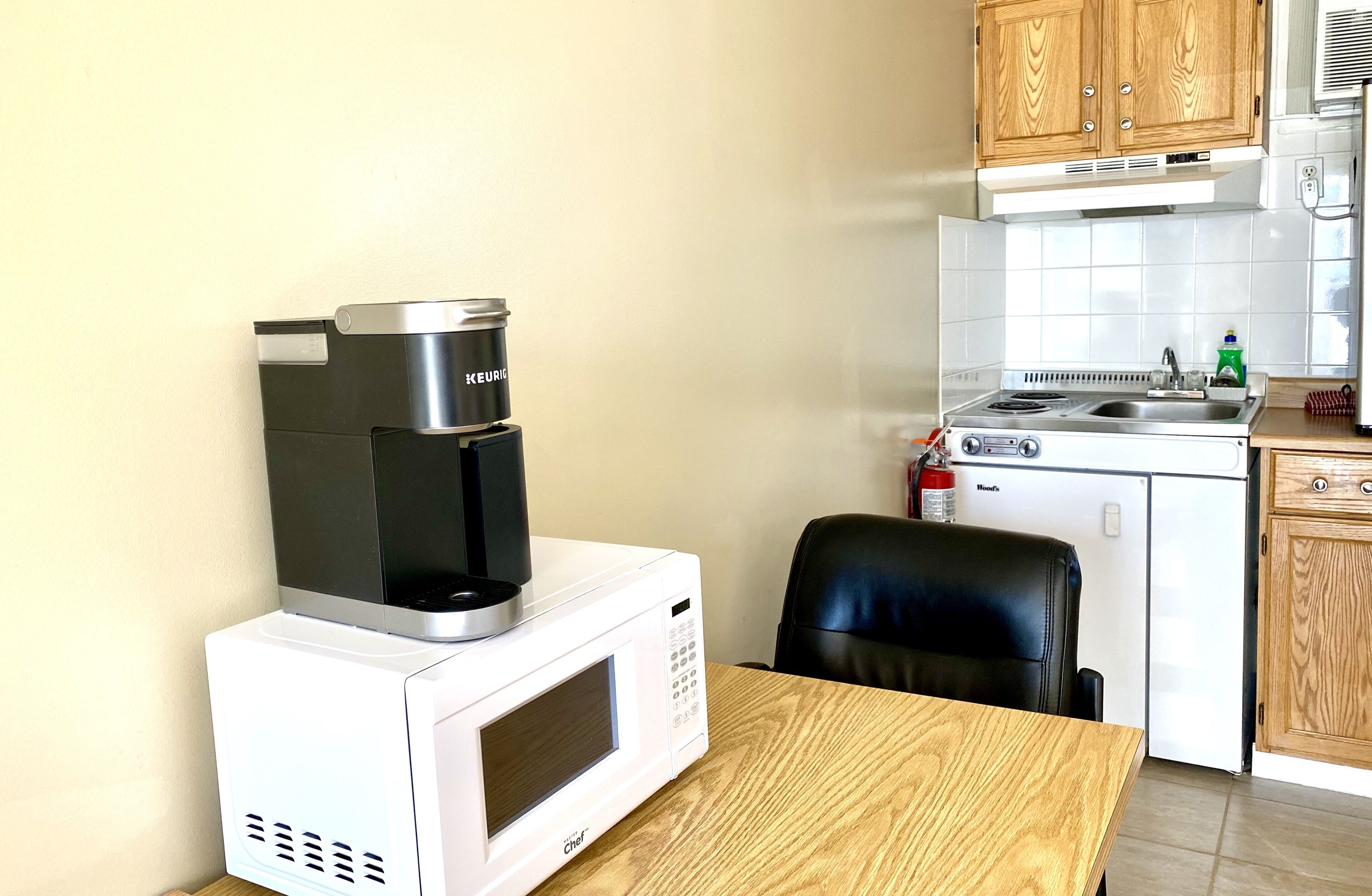 A compact kitchen with a microwave, coffee maker, and a small stove. There is a wooden table and a black office chair.