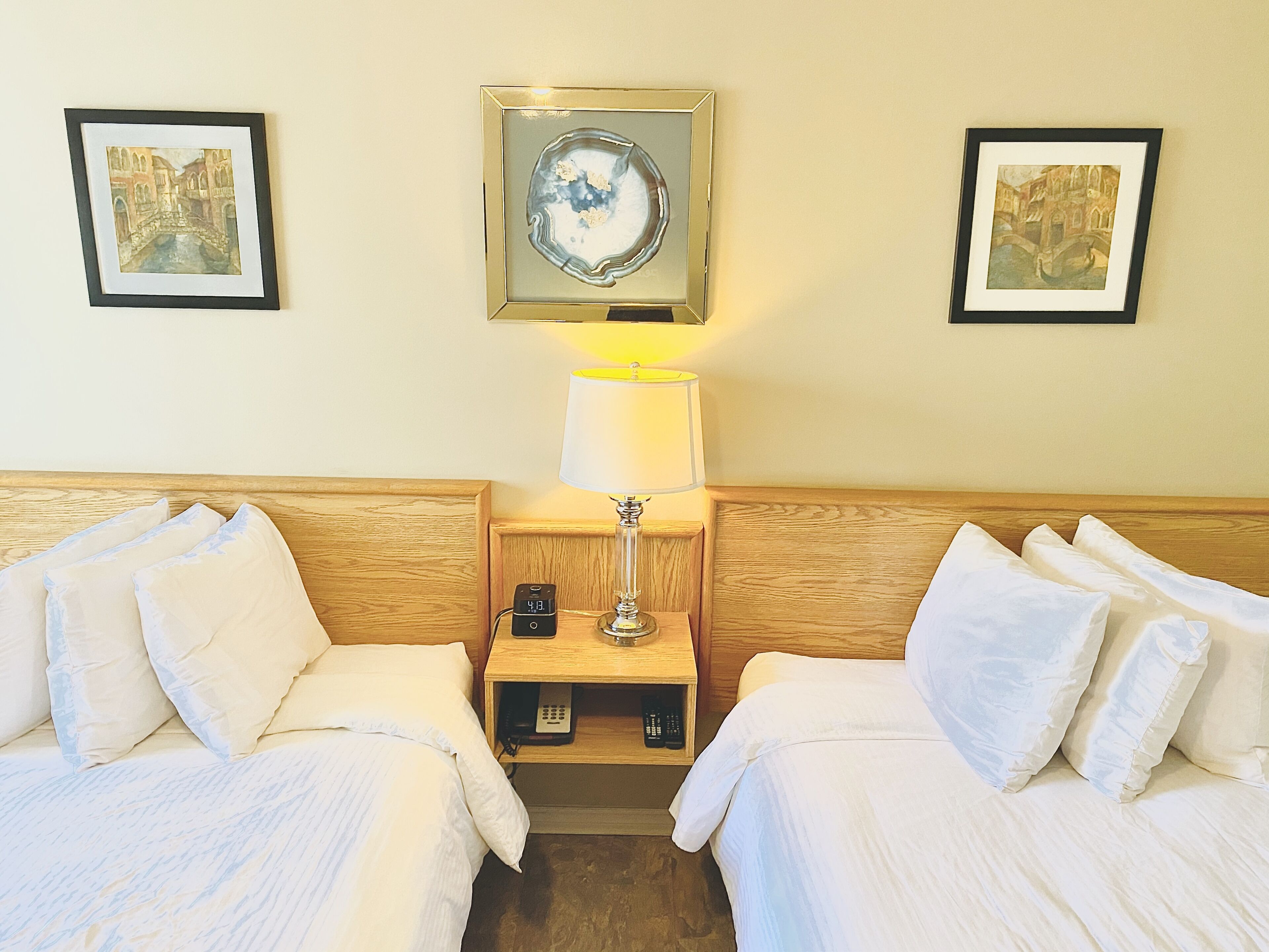 A hotel room with two beds, a nightstand with a lamp, a clock, and framed artwork on the walls.