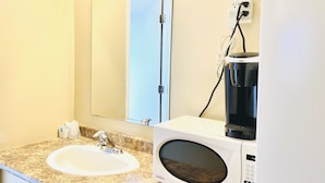 Un baño con lavamanos, espejo, microondas y cafetera.