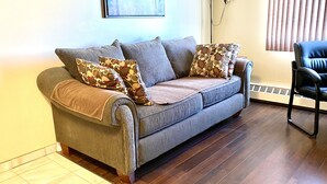 Un sofá gris con cojines estampados, una silla negra, una lámpara de pie y un radiador en una habitación con suelo de madera y una ventana con persianas.