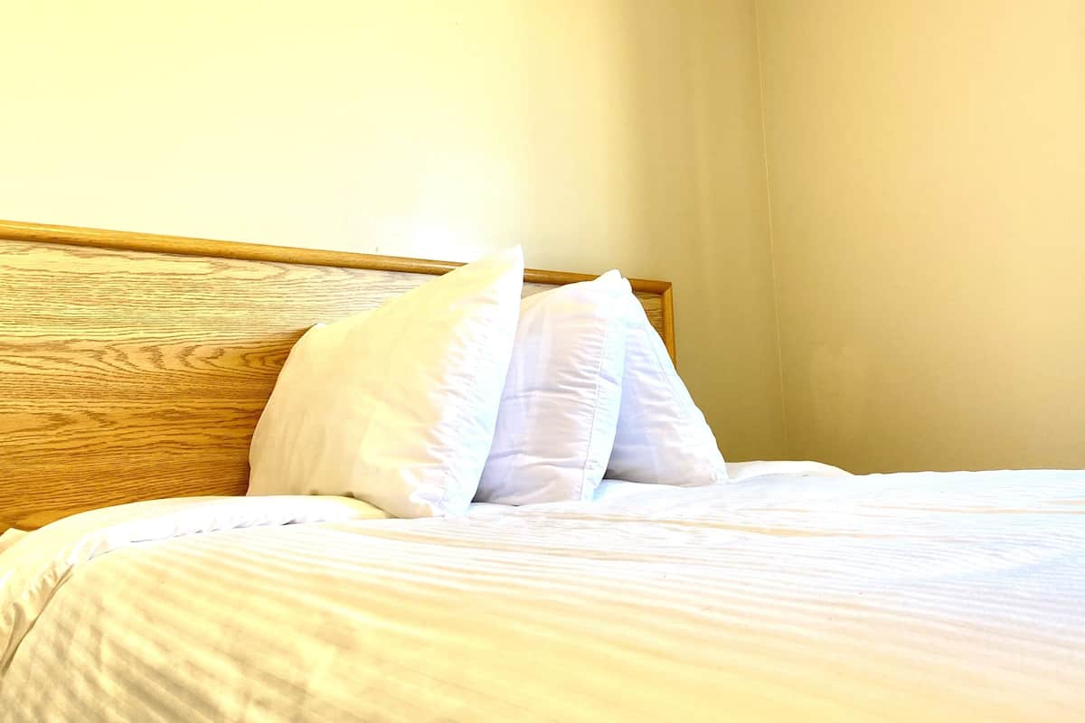 Ein Bett mit weißen Bettwäsche und Kissen, ein hölzernes Kopfteil und eine schlichte Wand.