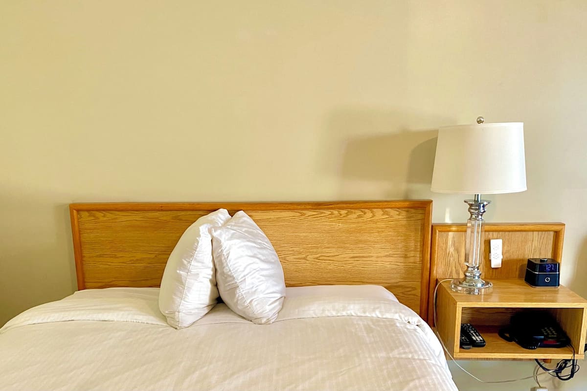 Ein ordentlich bezogenes Bett mit weißen Laken und zwei Kissen, eine Nachttischlampe, ein Nachttisch mit Lampe und digitaler Uhr sowie eine Fernbedienung.