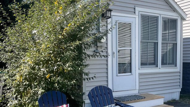 Une petite maison dotée d’un porche abrité, de deux fauteuils Adirondack avec des coussins aux couleurs du drapeau américain, et d’une terrasse en bois.