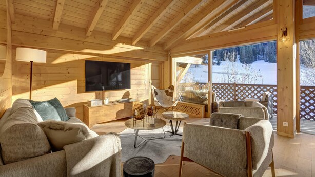 A cozy wooden cabin interior with a sofa, chairs, a TV, and a view of a snowy landscape. - Dorealp - superb apartment for 6 in peaceful setting - OVO Network (La Clusaz)