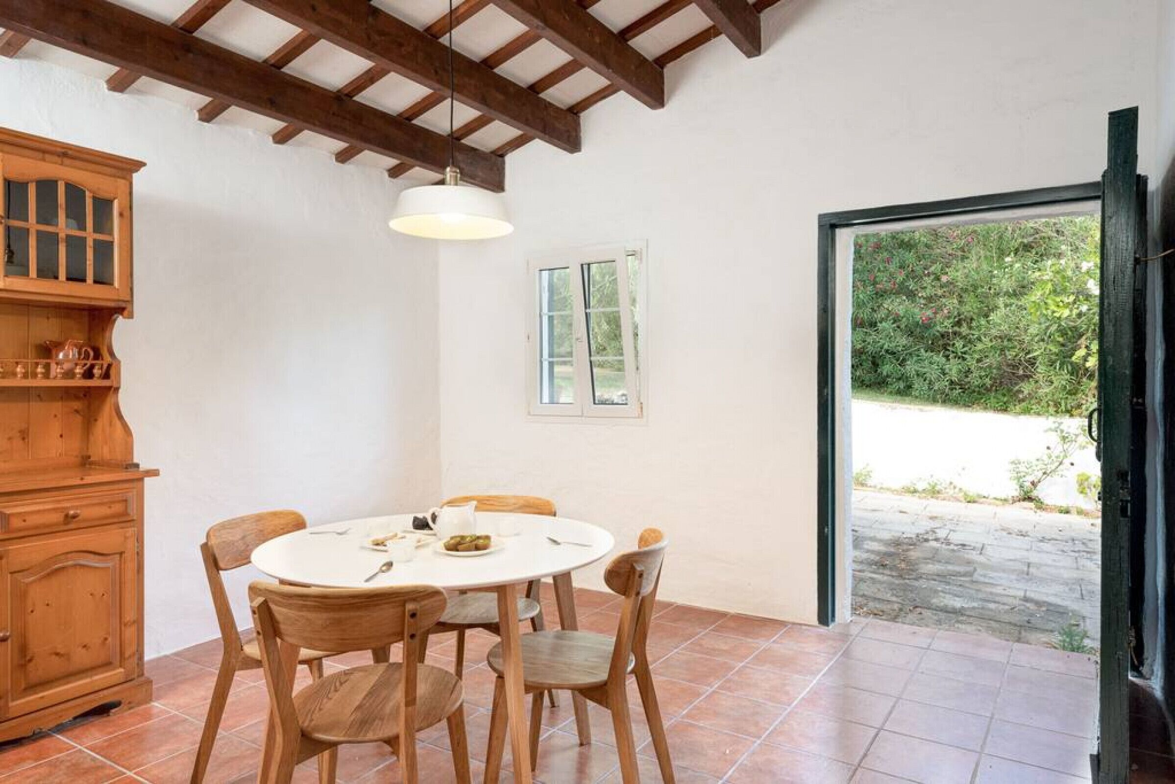 Una zona de menjador amb una taula rodona, cadires de fusta, un armari de fusta i una finestra que ofereix vistes a la vegetació.
