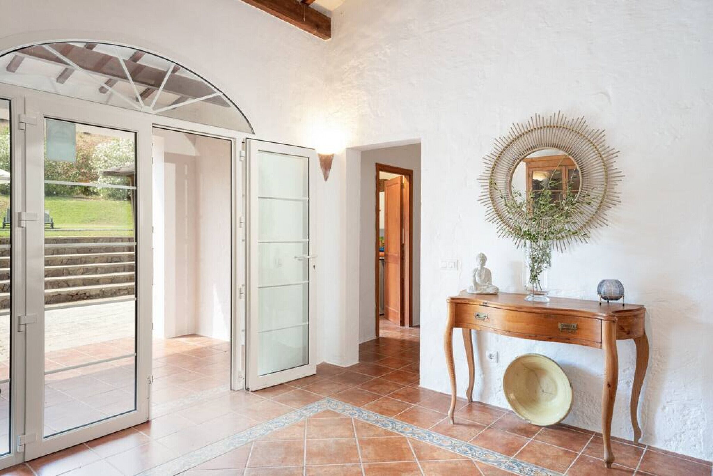 A hallway with a glass door leading to a patio, a wooden console table with a mirror, and a tiled floor.