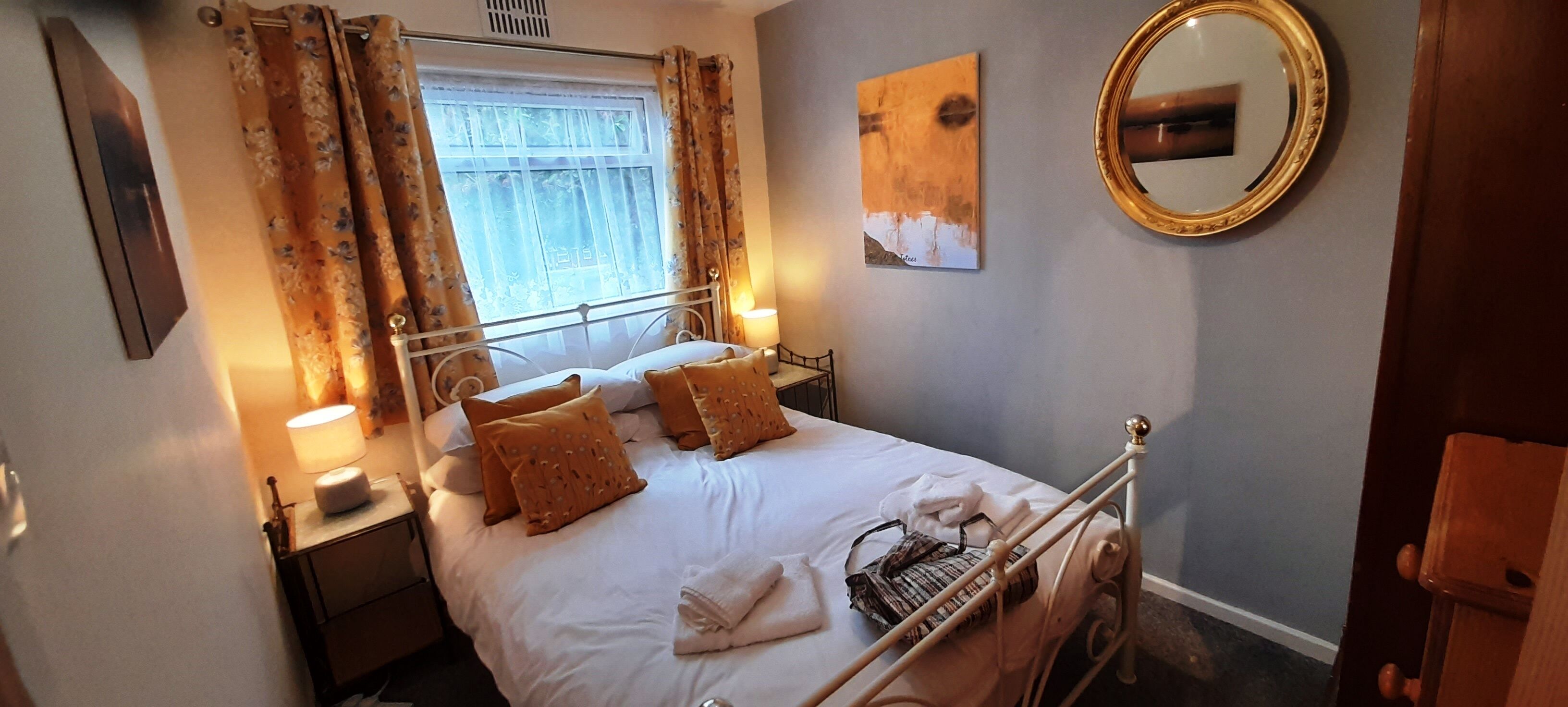 Een opgemaakt bed met witte lakens en twee bruine kussens, een bedlampje en een raam met gordijnen.