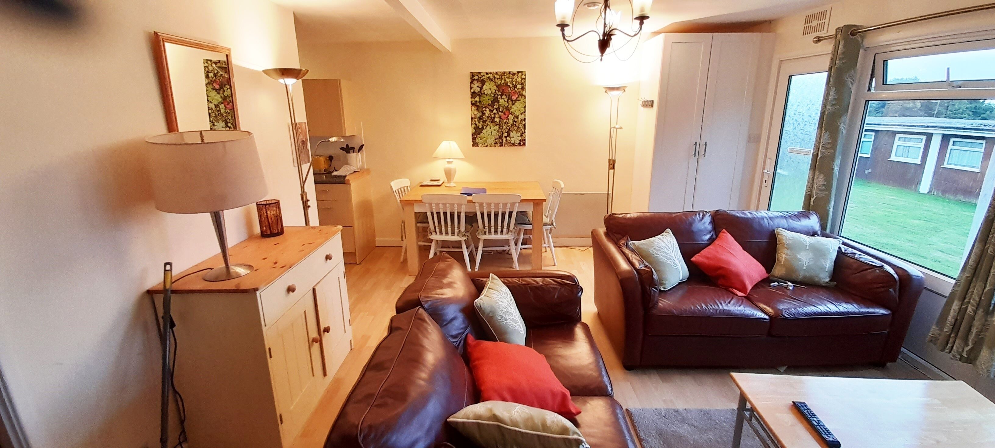 A living room with a brown leather sofa, a wooden dining table, and a kitchen area in the background.