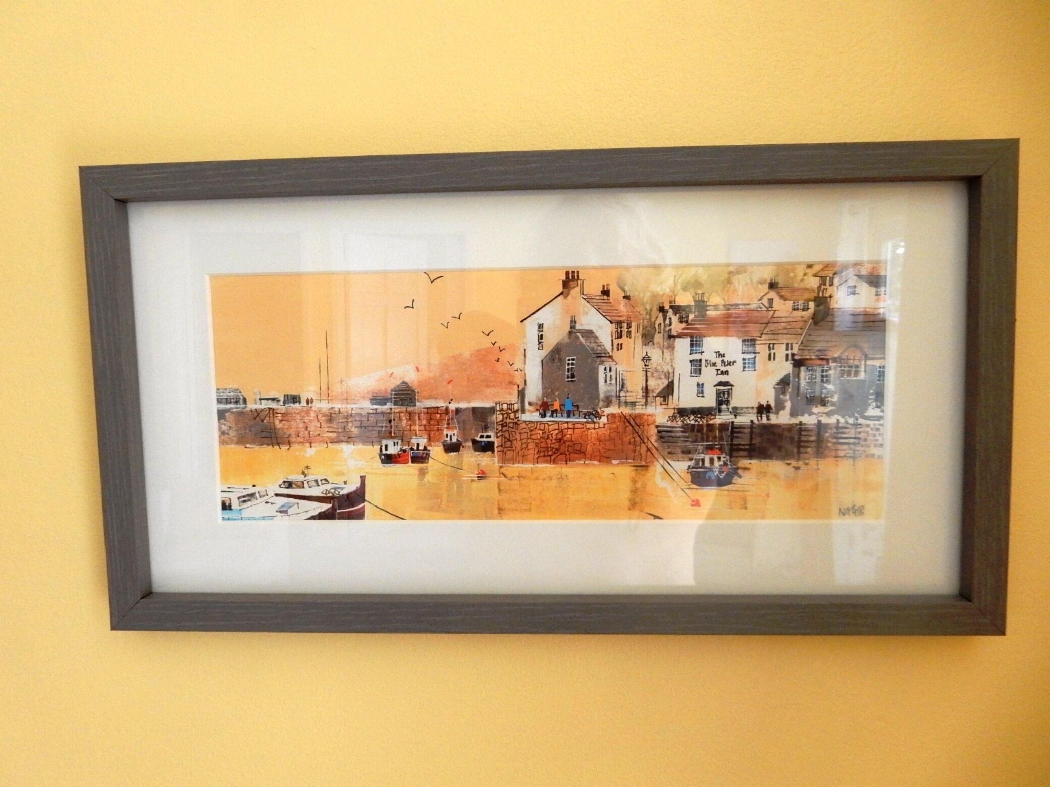 A framed artwork of a coastal town scene with buildings, boats, and people.