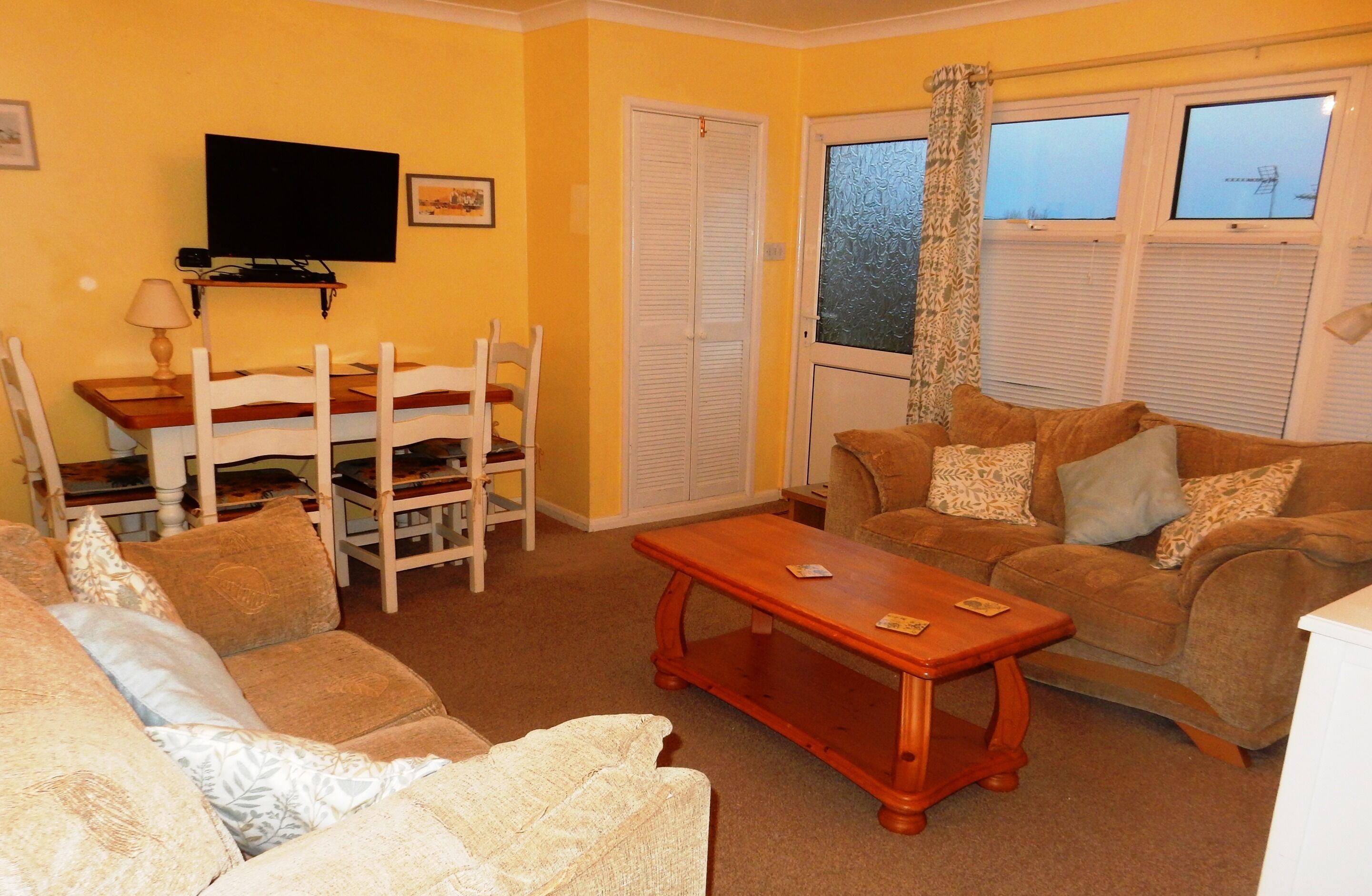 A living room with a sofa, a wooden coffee table, a TV mounted on the wall, a dining table with chairs, and a window with blinds.