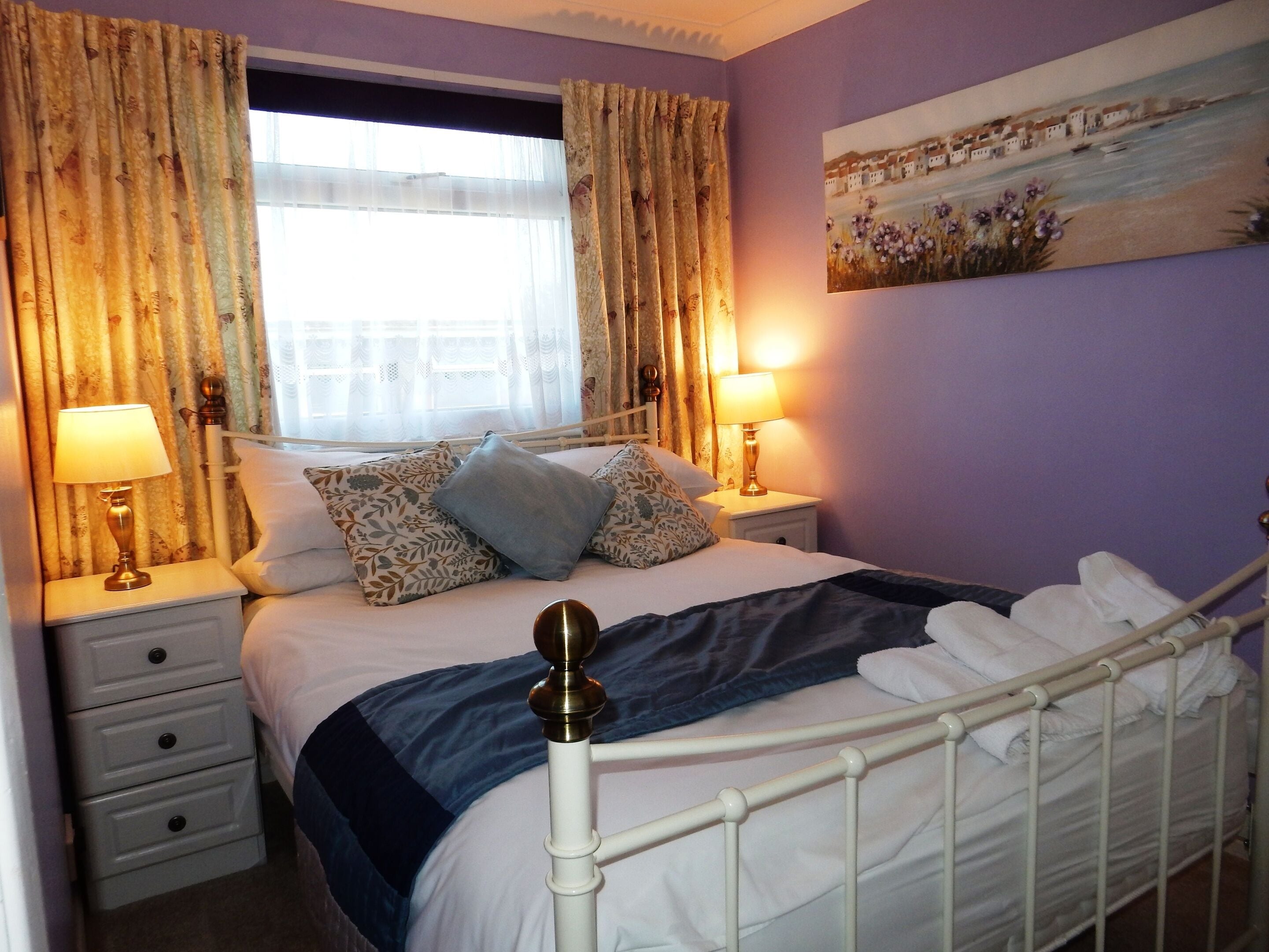 A neatly made bed with a blue blanket and pillows, two bedside lamps, a white nightstand, and a painting on the wall.