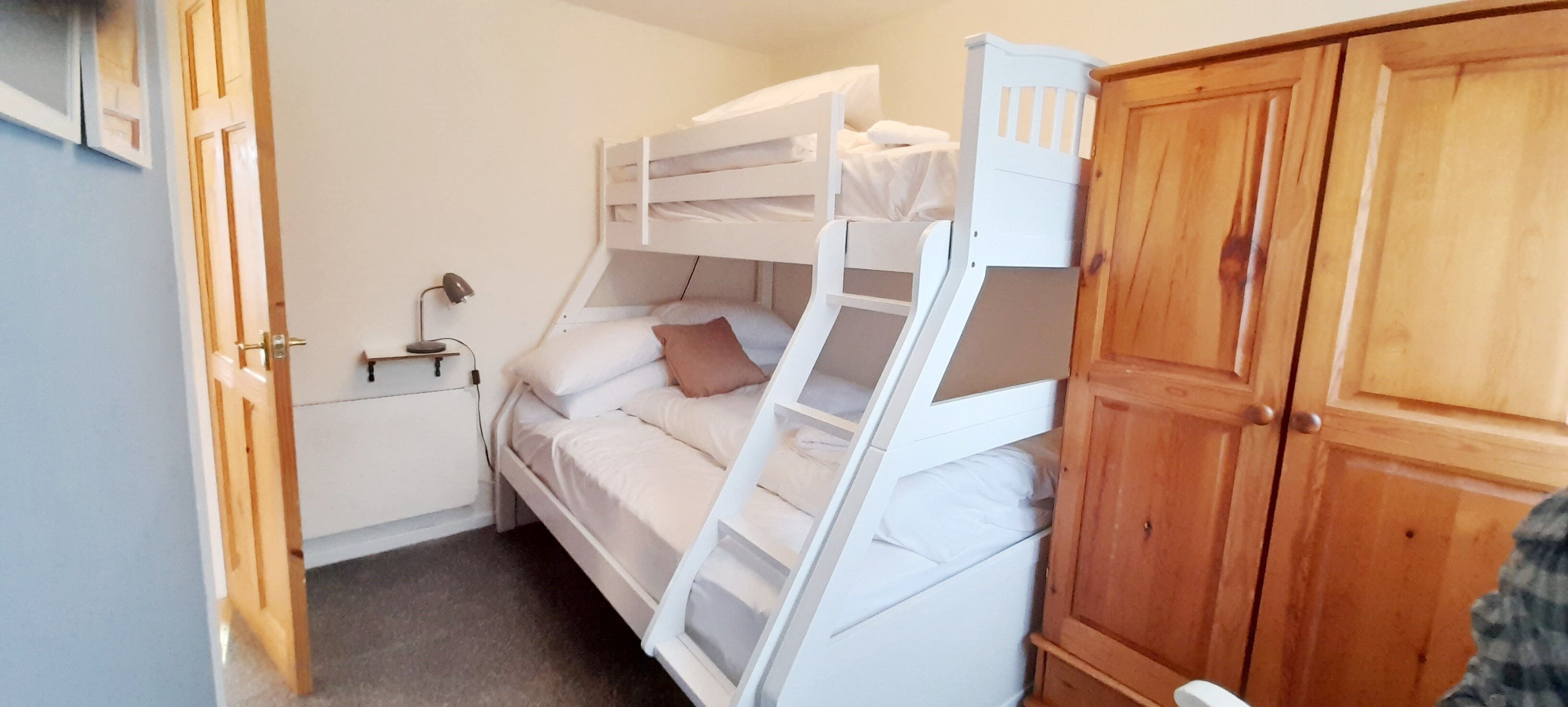 A bunk bed room with a wooden wardrobe and a desk with a lamp.