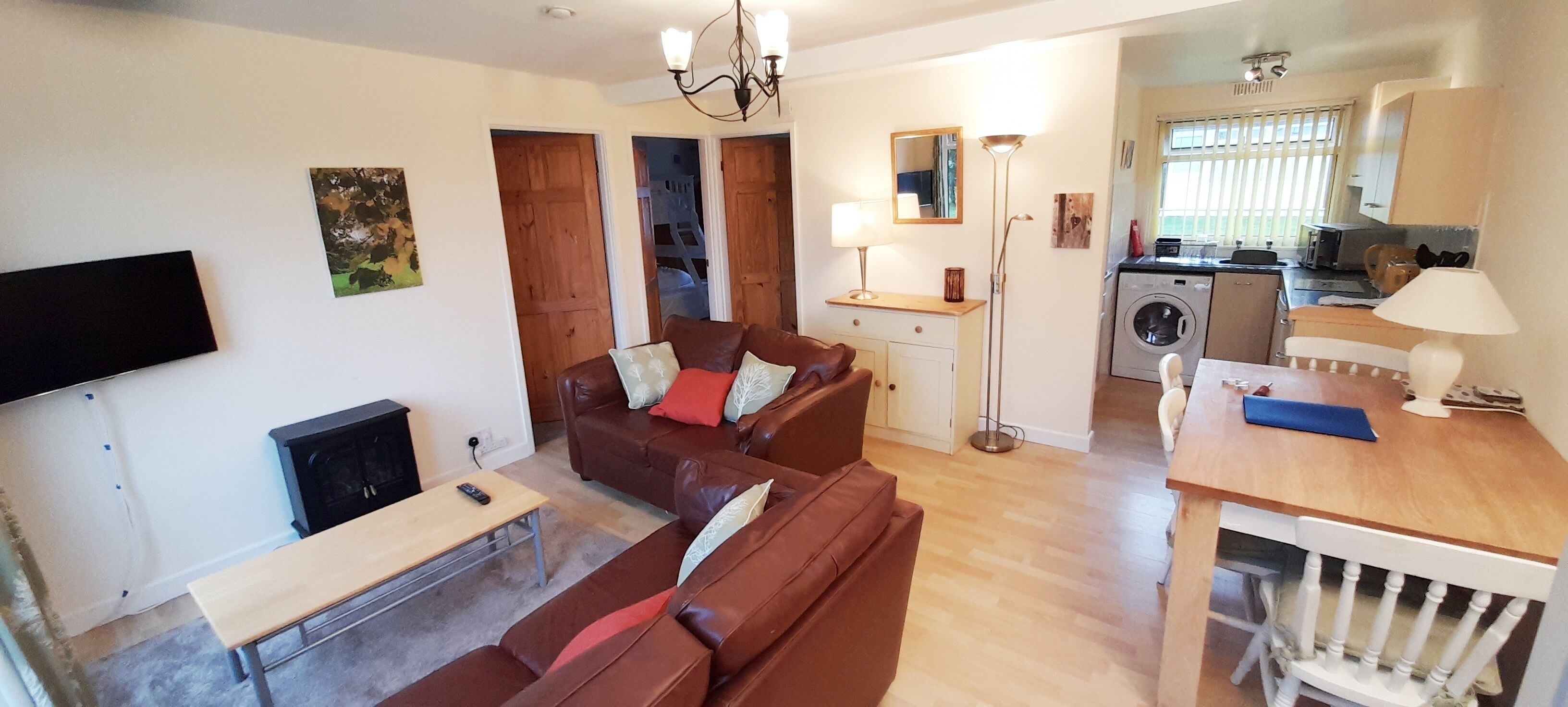 A living room with a brown leather sofa, a wooden coffee table, and a TV mounted on the wall. There is a kitchen area with a washing machine and a dining table with chairs.