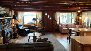 A cozy cabin interior with a stone fireplace, wooden furniture, and large windows offering a view of the outdoors. - Traverse City @ Rennie Lake  .. Idyllic Lakefront Retreat ..  CC49 ... Perfect! (traverse city)