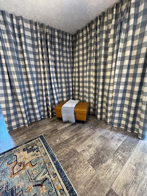 A room with checkered curtains, a wooden floor, a small orange ottoman with a blanket, and a patterned rug.