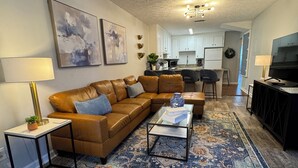 Ein Wohnzimmer mit braunem Ledersofa, einem Glastisch, einem gemusterten Teppich, einem Fernseher und einer Küche im Hintergrund.
