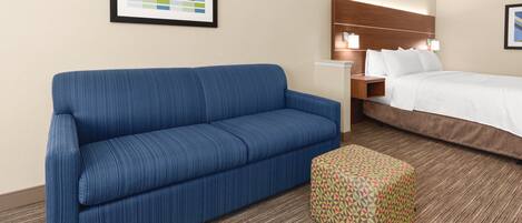 A hotel room with a blue sofa, a patterned ottoman, a bed with white bedding, a nightstand, and a framed picture on the wall.