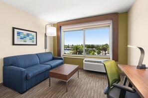 A hotel room with a sofa, a small table, a desk with a chair, a window with a view of the outdoors, and a wall-mounted lamp. - Holiday Inn Express & Suites Suisun City by IHG (Suisun City)