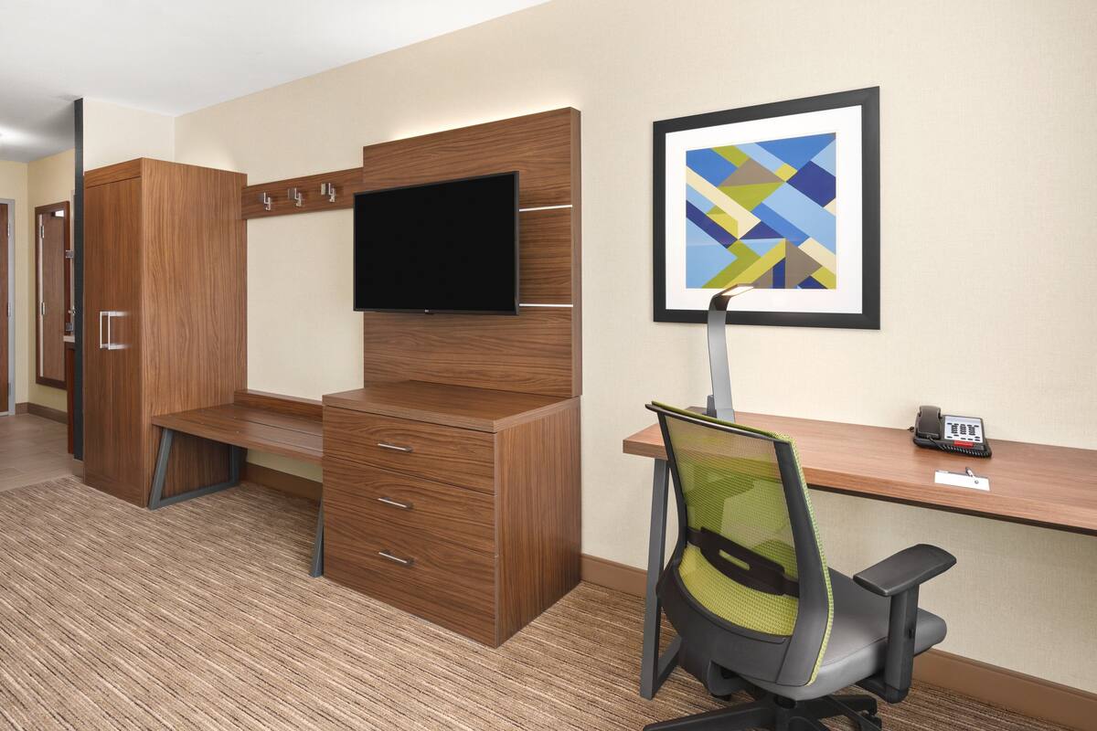 A hotel room with a wooden desk, a flat-screen TV, a green mesh office chair, and a framed abstract painting on the wall.