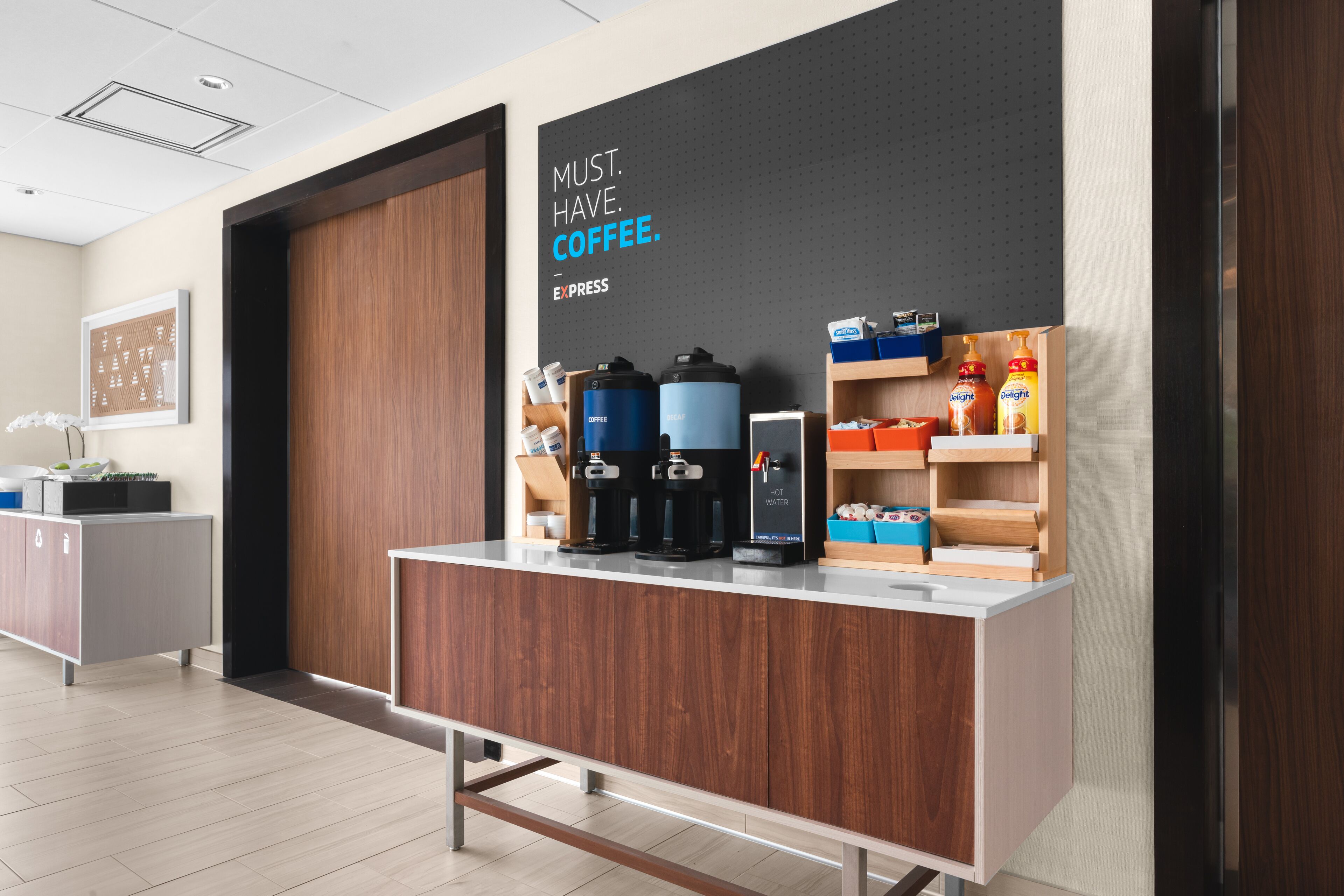 Un espace café moderne, agrémenté d’un panneau indiquant : « IL FAUT ABSOLUMENT AVOIR DU CAFÉ » et d’un distributeur d’eau.