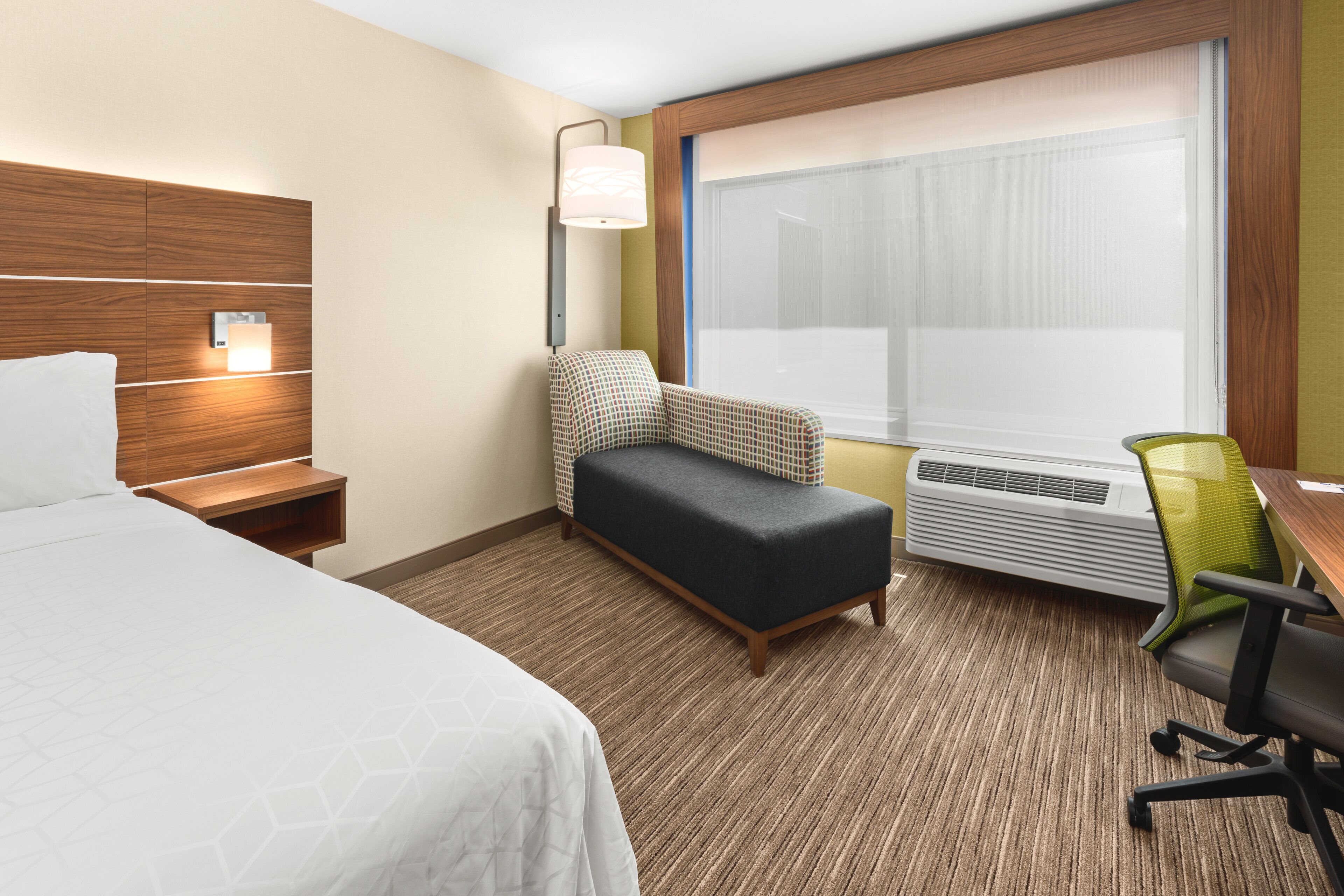 Habitación de hotel con cama, escritorio, silla, banco, lámpara de pared y ventana con persianas.
