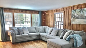 Ein L-förmiges Ecksofa mit vielen Kissen, große Fenster mit Vorhängen, Holzvertäfelung und ein Gemälde an der Wand.