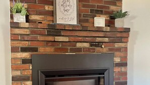 Ein Kamin aus Ziegelsteinen mit schwarzem Rahmen, darüber ein gerahmter Spruch und eine kleine Pflanze auf dem Sims.