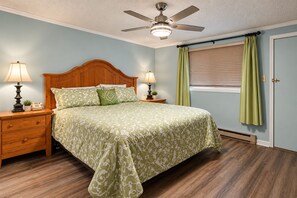 Ein Schlafzimmer mit einem Bett, zwei Nachttischen, Lampen, einem Deckenventilator, einem Fenster mit Jalousien und einer Tür.