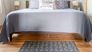 A neatly made bed with a grey bedspread and decorative pillows, a patterned rug on the wooden floor, and wall art above the bed.