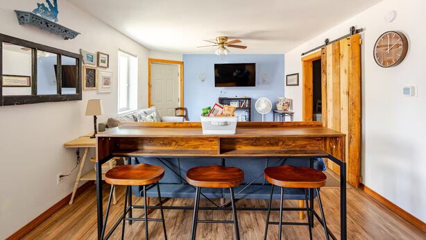 A kitchen with a wooden bar, three stools, a ceiling fan, a TV, and a clock on the wall.