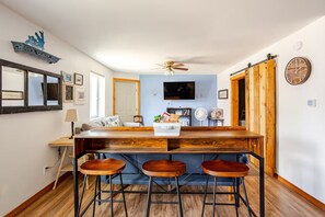 A kitchen with a wooden bar, three stools, a ceiling fan, a TV, and a clock on the wall. - Cozy Cottage near downtown brings Georgetown Lake & Fairmont to your fingertips! (Anaconda)