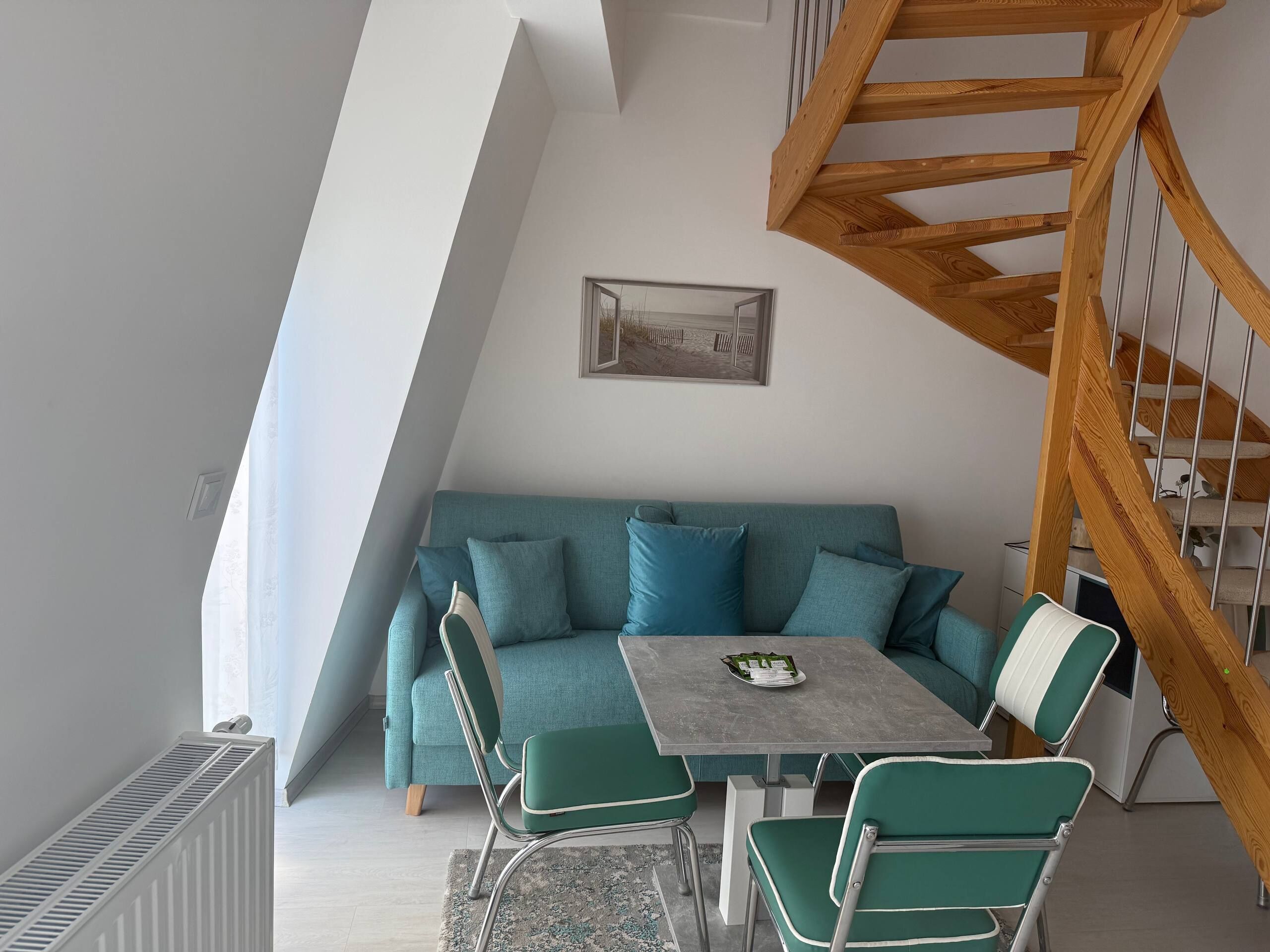 Een moderne loft slaapkamer met schuin dak, een blauwgroene bank, een eettafel en een houten trap.