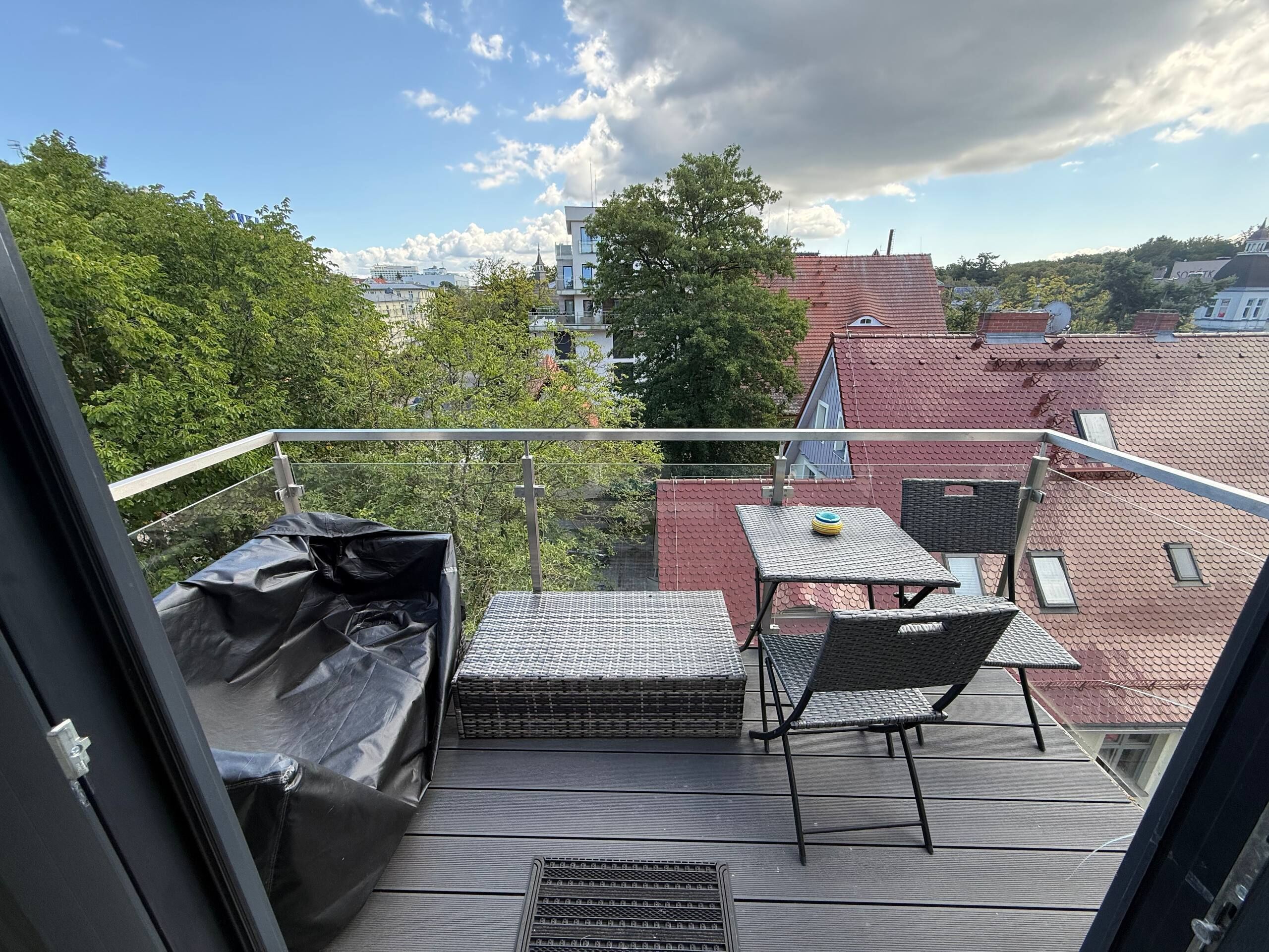 Een balkon met een tafel en stoelen, een overdekte zithoek, en uitzicht op gebouwen en bomen.