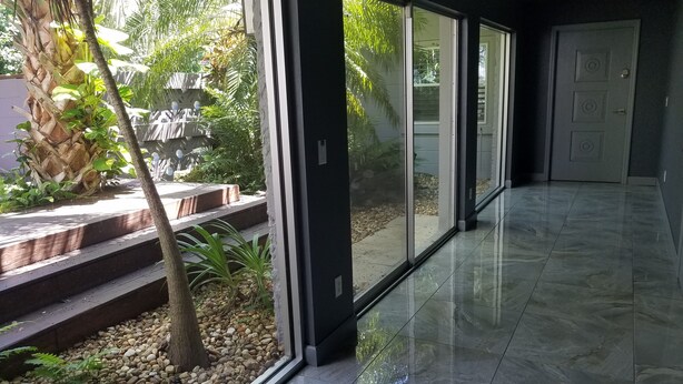 Un couloir moderne doté de grandes portes vitrées donnant sur un jardin, où l’on peut admirer un palmier et une petite cascade.