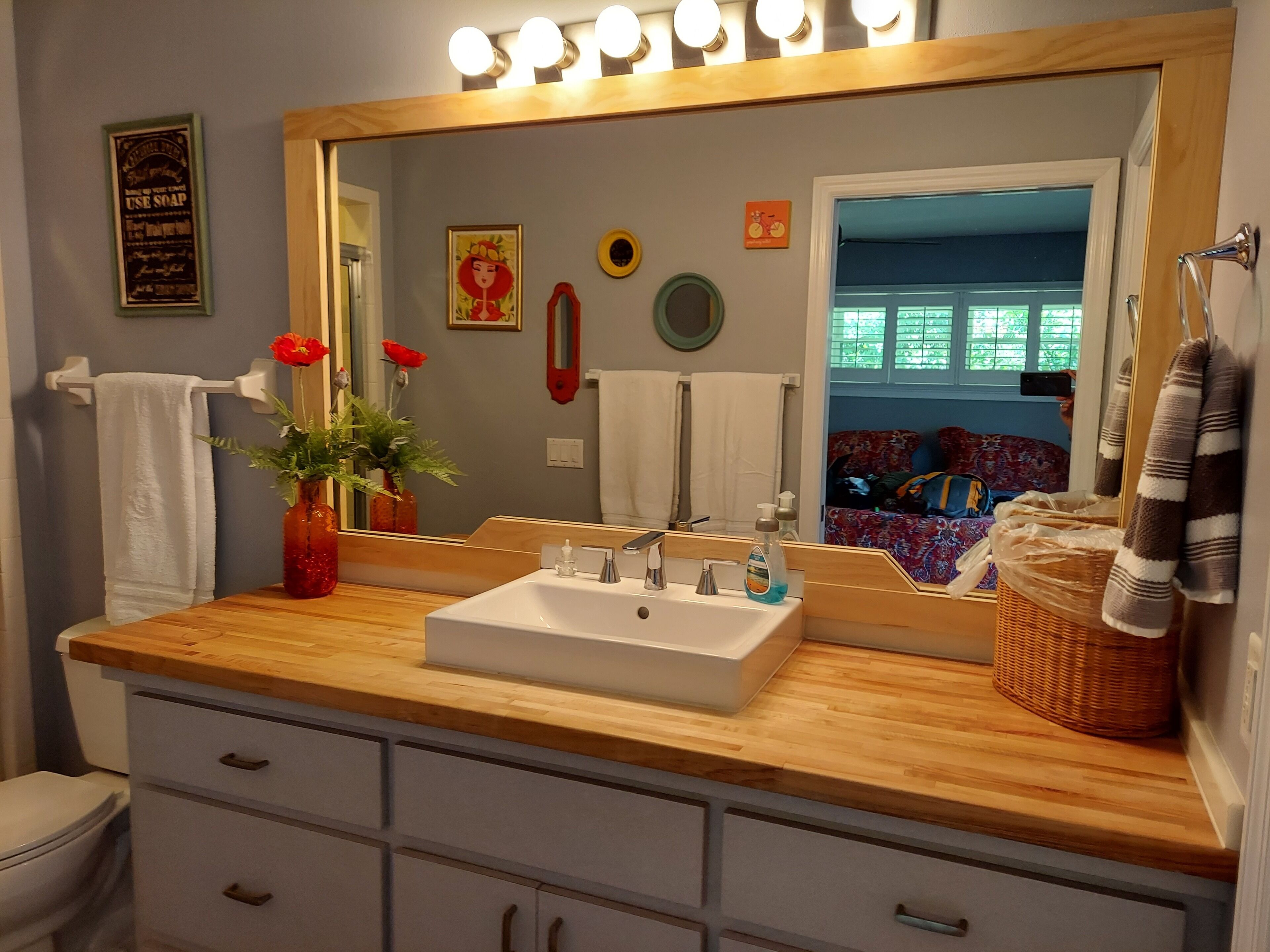 A bathroom with a wooden vanity, white sink, and a large mirror with lights. There are towels, a vase with flowers, and a basket on the counter.