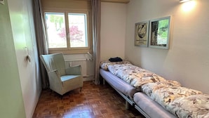 Een kleine kamer met een houten parketvloer, een enkel bed met bloemenbeddengoed, een stoel, een radiator en een raam met gordijnen.