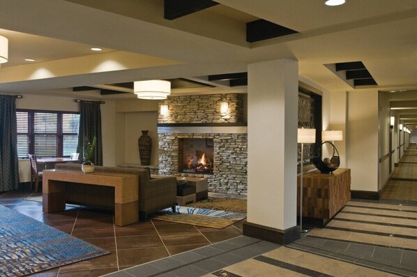 A modern hotel lobby with a stone fireplace, a seating area with a sofa and a coffee table, and a hallway with a patterned rug.