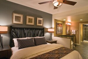 Ein Hotelzimmer mit einem großen Bett, einem Deckenventilator, gerahmten Kunstwerken an der Wand, einem Badezimmer mit Dusche und einem kleinen Schreibtisch mit Lampe.