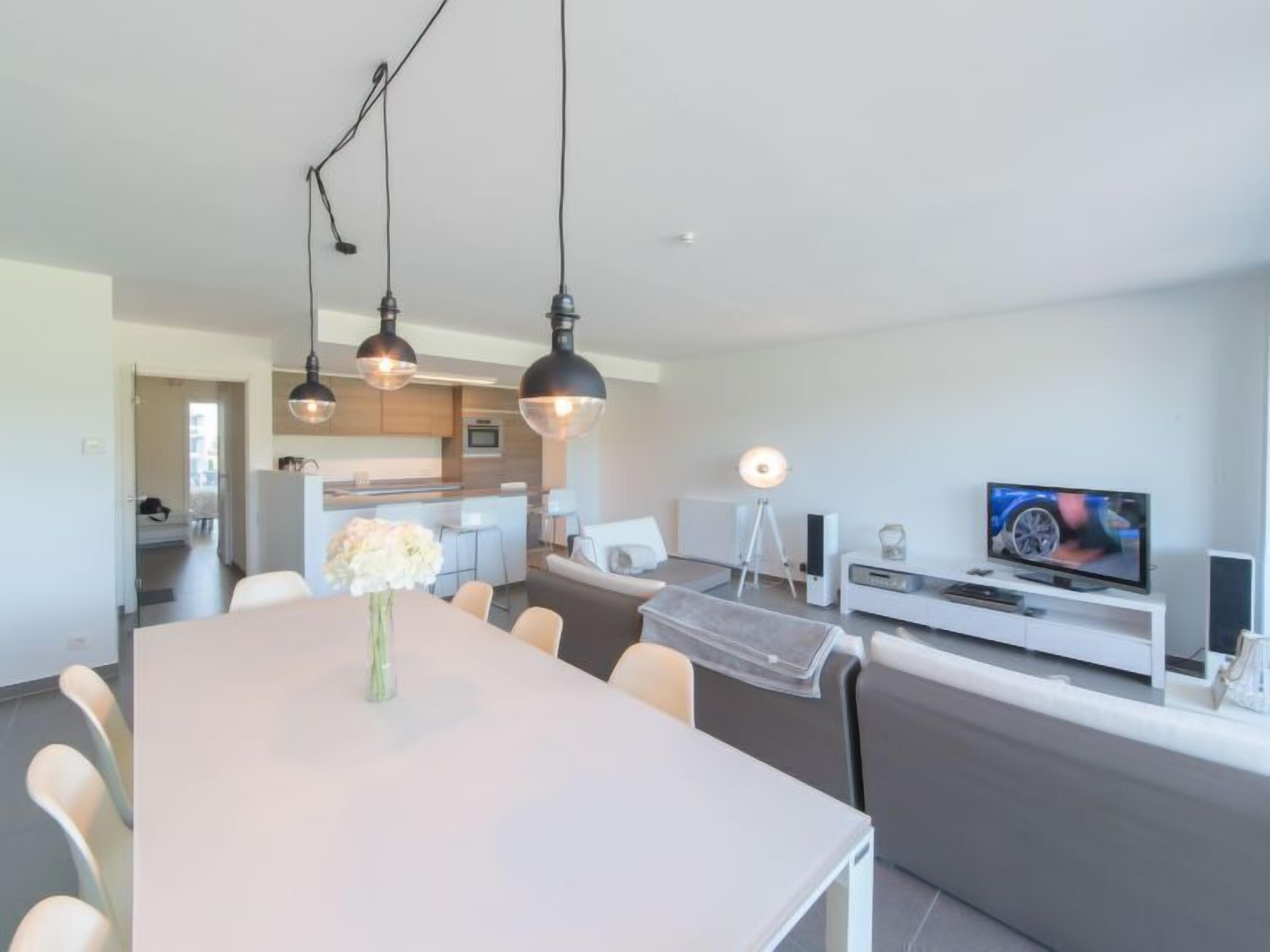 A modern living room with a dining area, a kitchen in the background, and a TV on a white console.